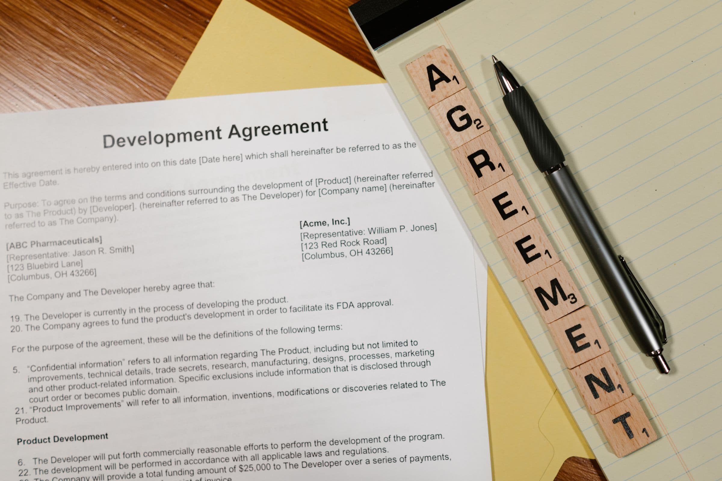 机の上に置かれた開発契約書とボールペン、AGREEMENTの文字を並べた木製タイル
